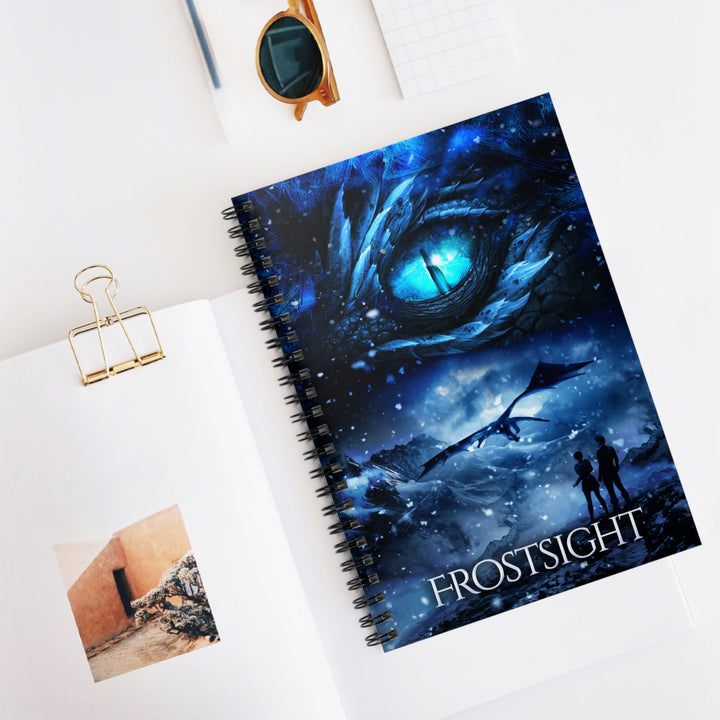 A notebook with a cover featuring the artwork from the cover of Frostsight. Artwork features a blue dragon eye at the top. Below the eye, the two main characters watch a dragon in the distance in a snowy mountain landscape. The title is at the bottom of the notebook cover.