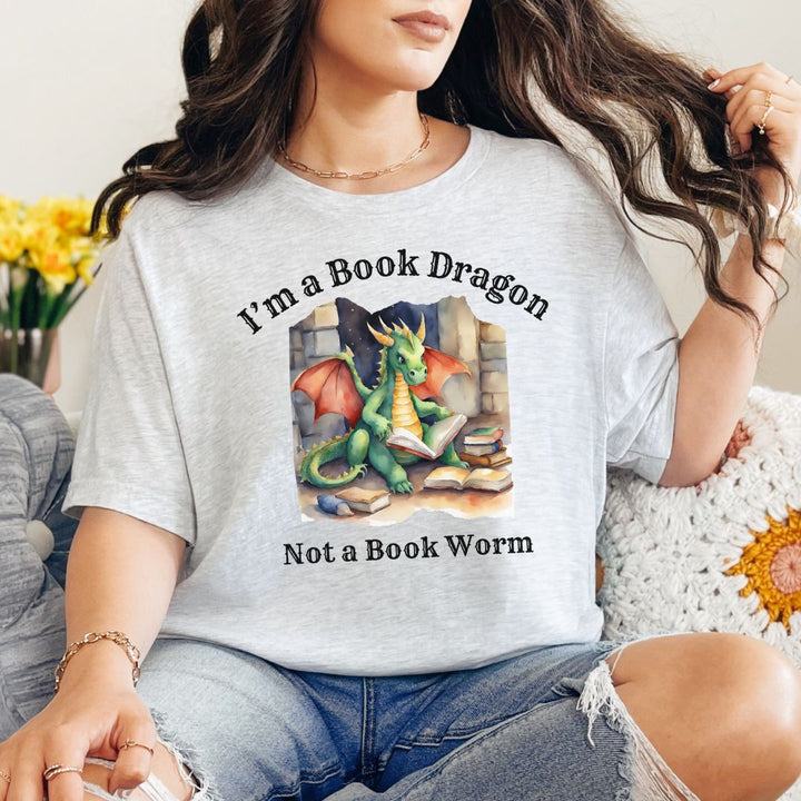 A woman wearing a short sleeve heather gray t-shirt. Design on the shirt reads "I'm a book dragon not a book worm." Between the text is a watercolor design of a dragon reading next to a stack of books.