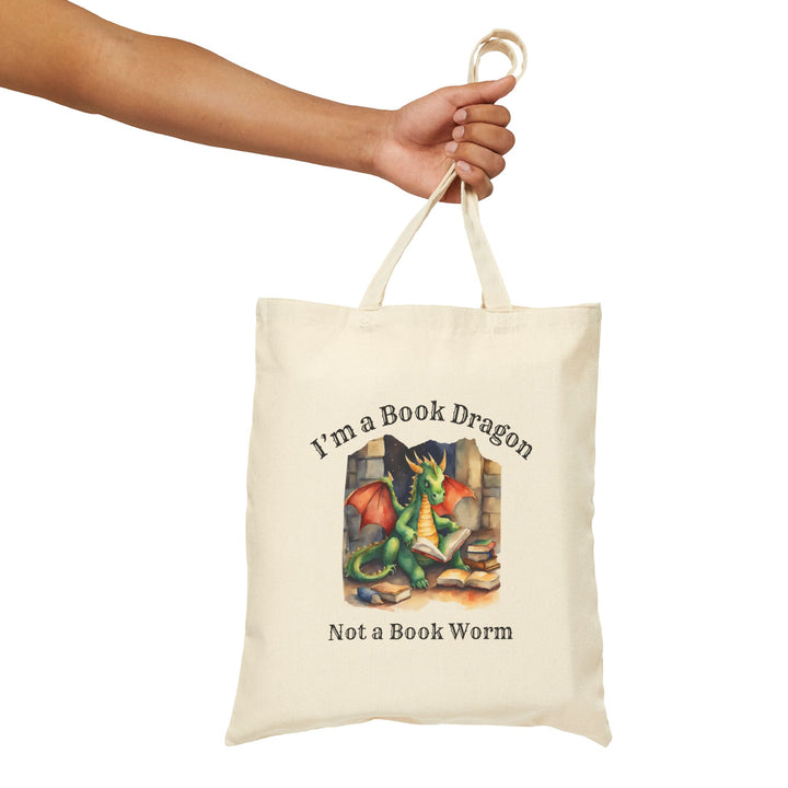 Someone holding a canvas tote bag. Design on the bag reads "I'm a book dragon not a book worm." Between the text is a watercolor design of a dragon reading next to a stack of books.