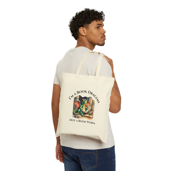 Someone holding a tan canvas tote bag. Design on the tote bag reads "I'm a book dragon not a book worm." Between the text is a watercolor design of a dragon reading next to a stack of books.