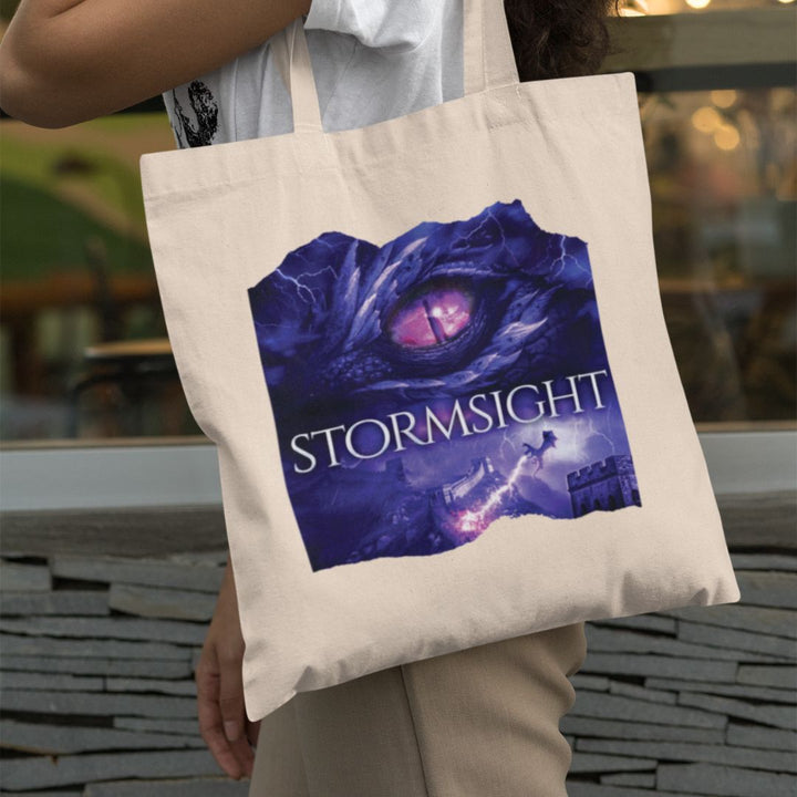 A person holding a tote bag. Design on the bag is the cover of Stormsight, just showing the purple dragon eye and the title with the a dragon breathing lightning onto a wall.