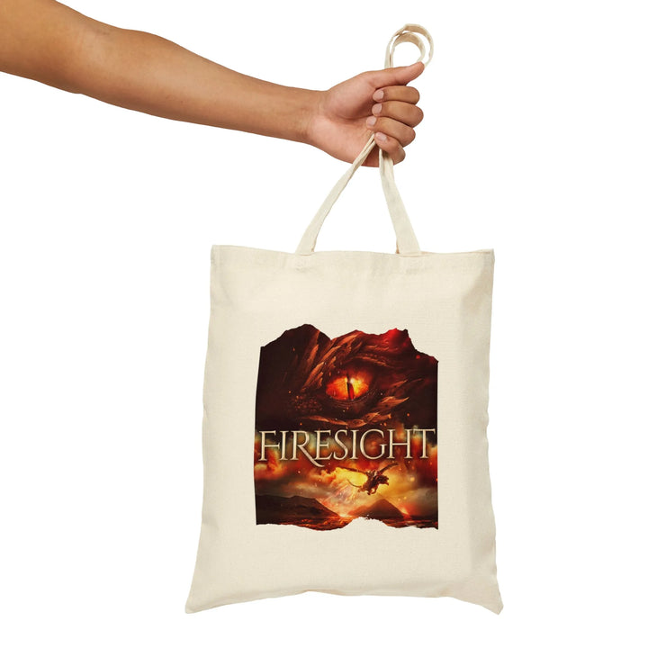Someone holding a tan canvas tote bag. Design on the tote bag is the cover of Firesight, just showing the red dragon eye and the title with the two main characters riding on a dragon over a volcano erupting.