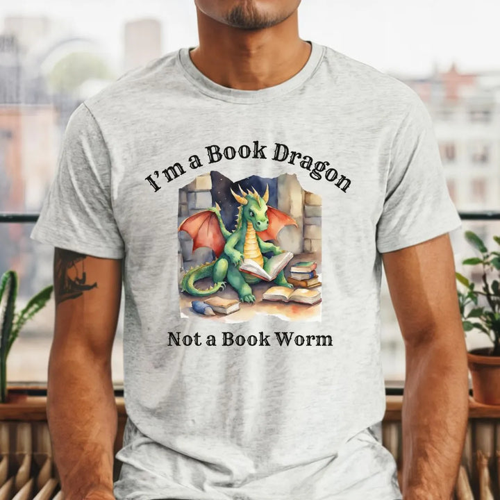 A man wearing a short sleeve heather gray t-shirt. Design on the shirt reads "I'm a book dragon not a book worm." Between the text is a watercolor design of a dragon reading next to a stack of books.