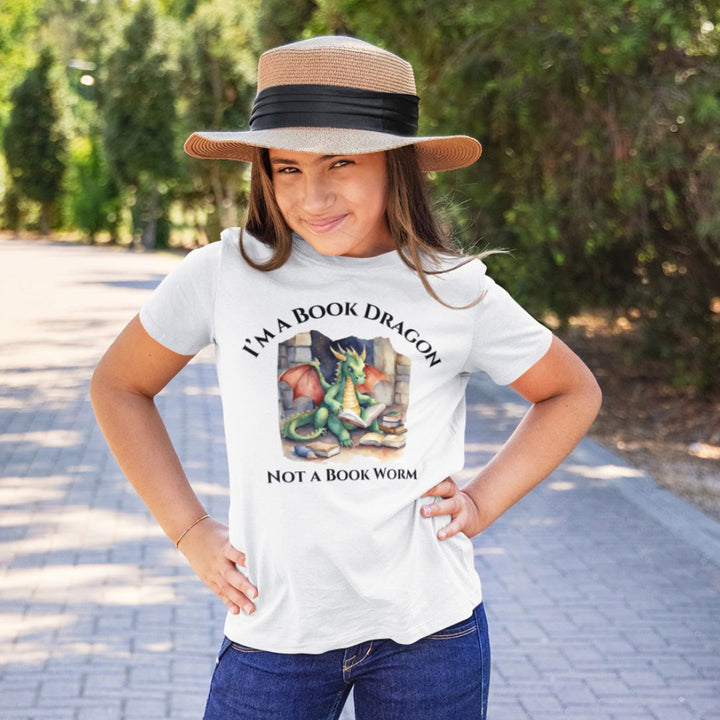 A young girl wearing a short sleeve heather gray t-shirt. Design on the shirt reads "I'm a book dragon not a book worm." Between the text is a watercolor design of a dragon reading next to a stack of books.