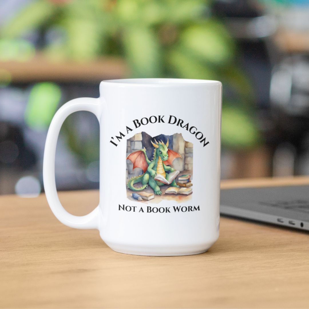 A white mug sitting on a desk. Design on the mug reads "I'm a book dragon not a book worm." Between the text is a watercolor design of a dragon reading next to a stack of books.
