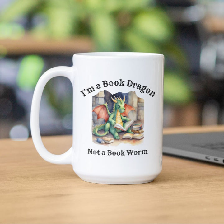 A white mug sitting on a table. Design on the mug reads "I'm a book dragon not a book worm." Between the text is a watercolor design of a dragon reading next to a stack of books.