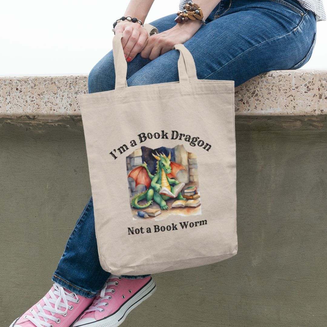 Someone holding a canvas tote bag. Design on the bag reads "I'm a book dragon not a book worm." Between the text is a watercolor design of a dragon reading next to a stack of books.