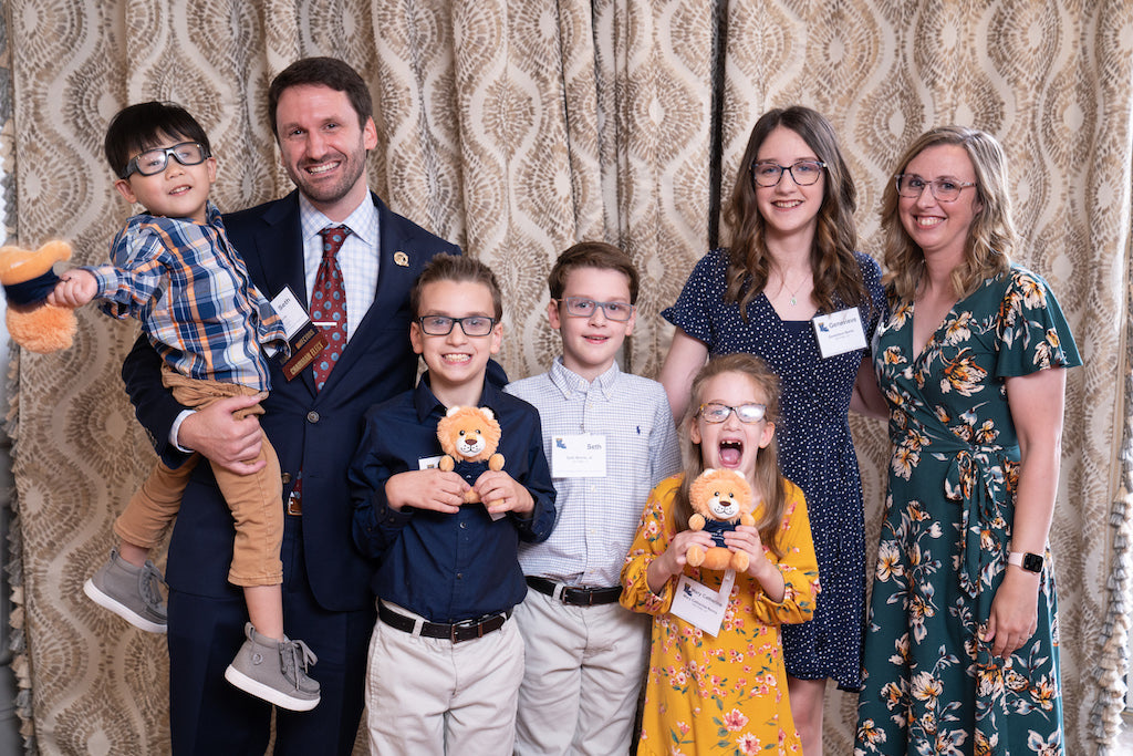 The author Jessica Deen Norris, her husband, and her five children smile for a photo. They are dressed up for her husband's business event. Her three youngest children are holding little stuffed lion toys and her youngest daughter has a big silly smile.