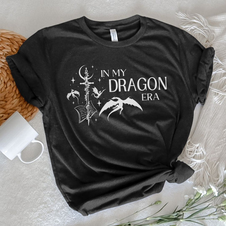 A short sleeve heather black t-shirt. The white design on the shirt reads "In my dragon era." Next to the text are a sword, a book, magical stars and moon, and flying dragons.