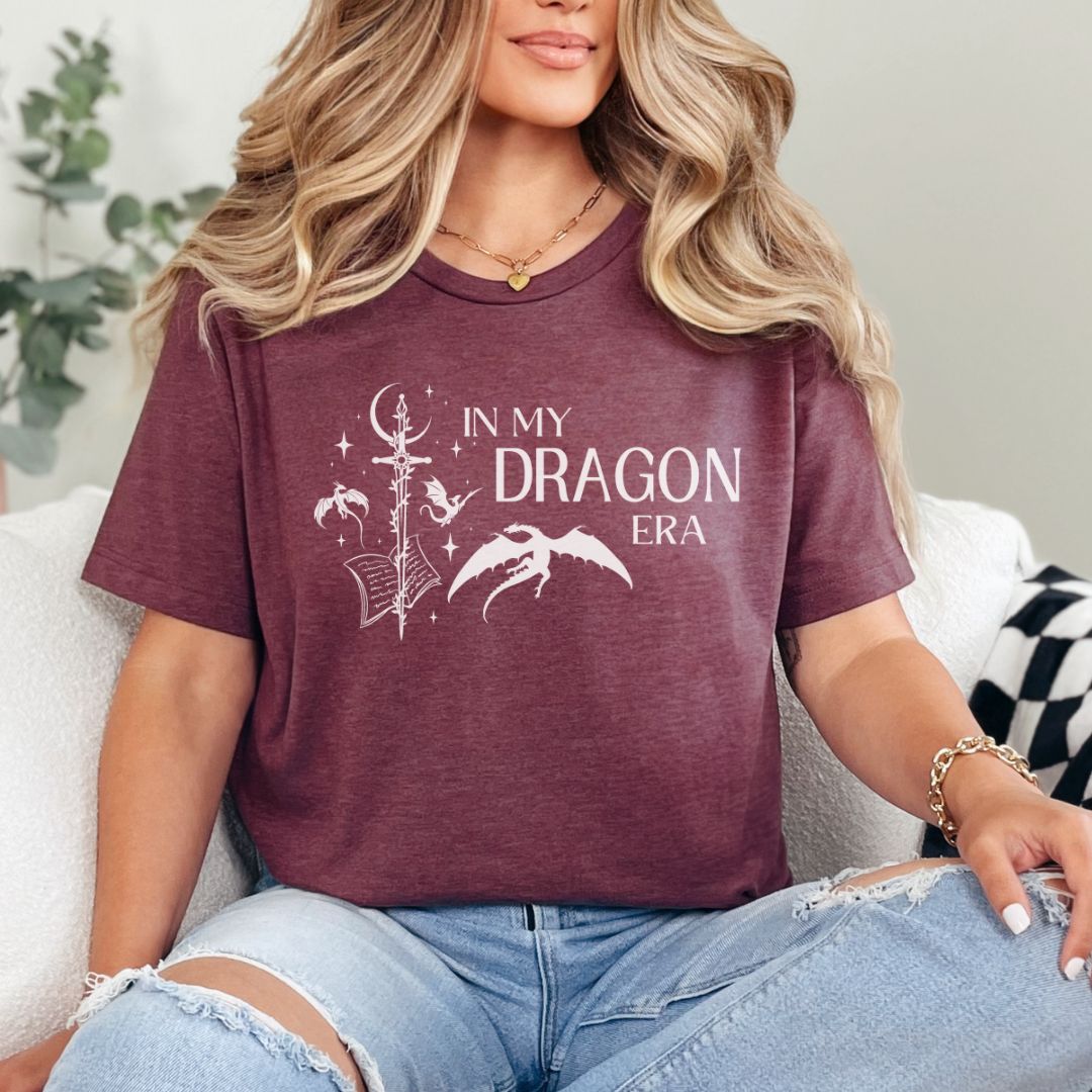 A woman wearing a short sleeve heather maroon t-shirt. The white design on the shirt reads "In my dragon era." Next to the text are a sword, a book, magical stars and moon, and flying dragons.