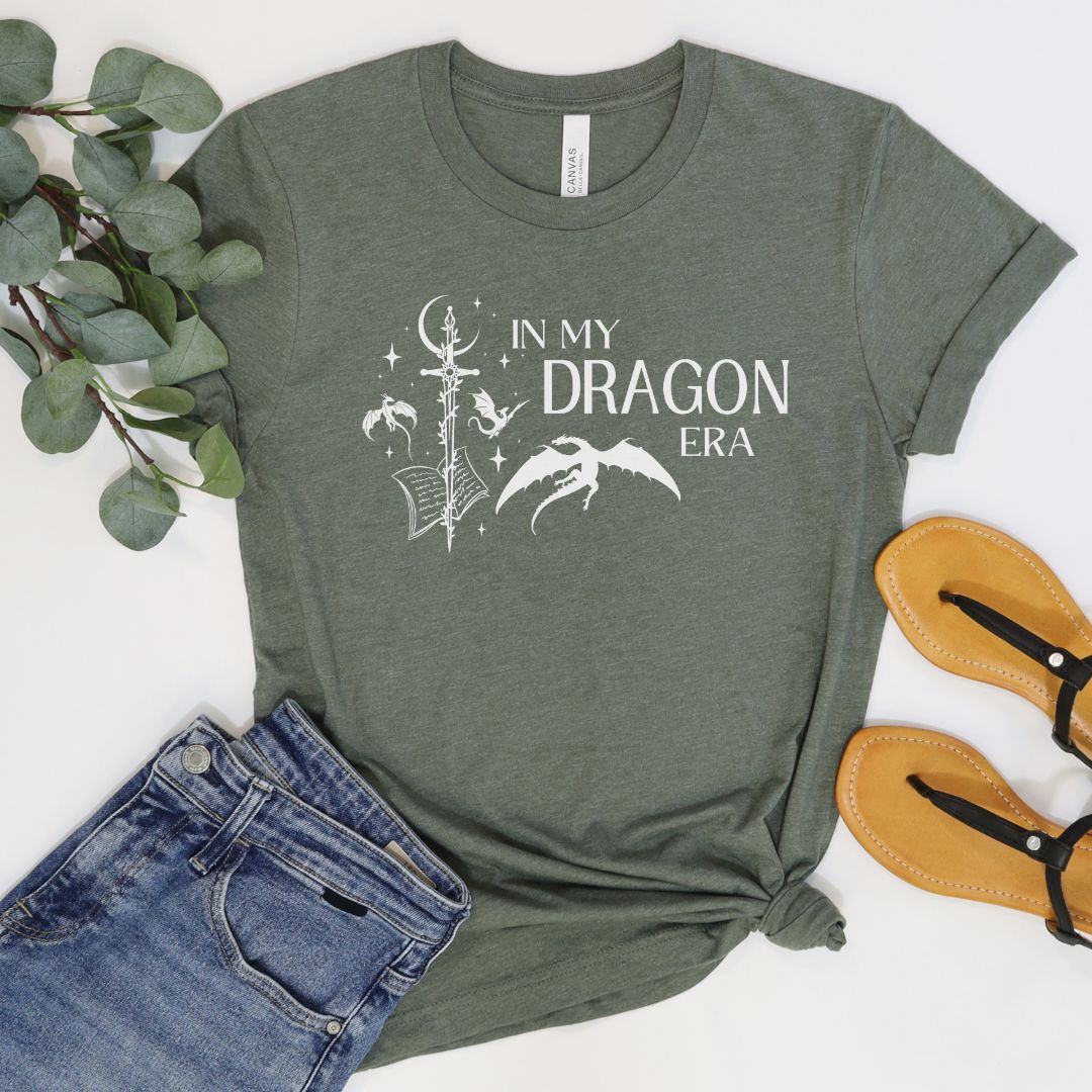 A short sleeve heather military green t-shirt. The white design on the shirt reads "In my dragon era." Next to the text are a sword, a book, magical stars and moon, and flying dragons.