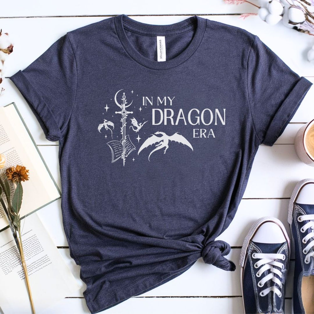 A short sleeve heather navy t-shirt. The white design on the shirt reads "In my dragon era." Next to the text are a sword, a book, magical stars and moon, and flying dragons.