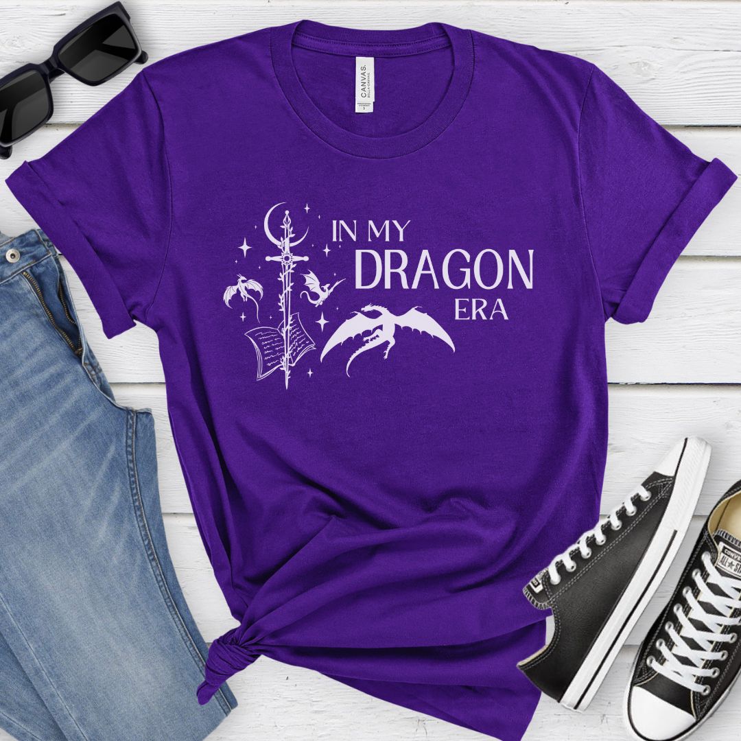 A short sleeve purple t-shirt. The white design on the shirt reads "In my dragon era." Next to the text are a sword, a book, magical stars and moon, and flying dragons.