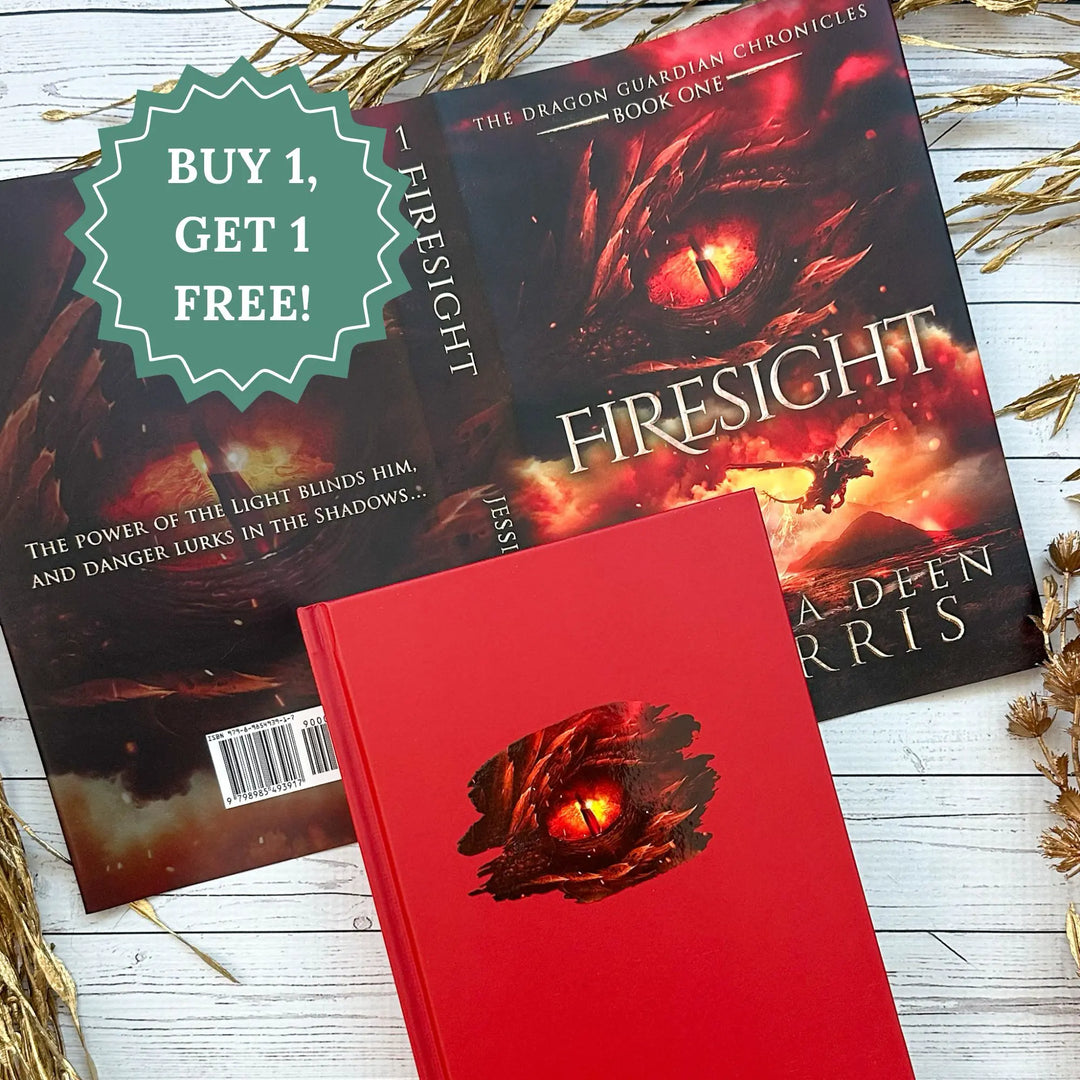 A red hardcover book titled 'Firesight' by Jessica Deen Norris is displayed in front of an open copy of the same book, which shows a dramatic red and orange dragon-eye cover. A green promotional badge reading 'BUY 1, GET 1 FREE!' is placed on top, with dried grasses on a white wood surface.