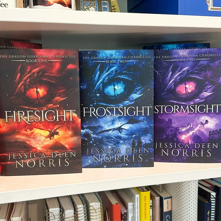 Three hardcover books from The Dragon Guardian Chronicles series by Jessica Deen Norris are displayed on a white bookshelf. The books are &