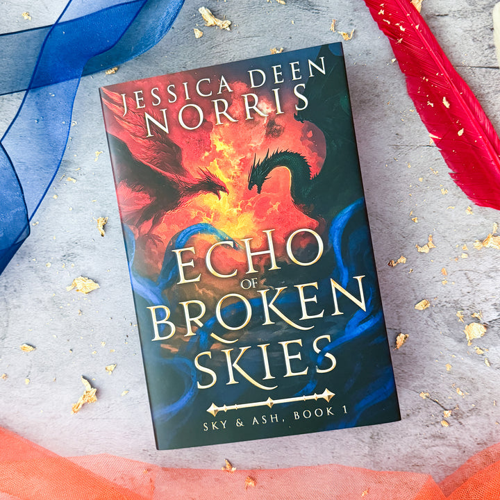 Hardcover - Echo of Broken Skies by Jessica Deen Norris - Cover features a sky divided by swirling clouds of fire. On the left, a red phoenix with outstretched wings flies toward a dark green dragon on the right, both poised as if mid-battle. Blue, smoky tendrils swirl around the bottom of the image. The title is centered in metallic gold, with Sky & Ash, Book 1 below a decorative gold divider. Sprayed edges feature the dragon and phoenix.#autograph-option_signed-by-author