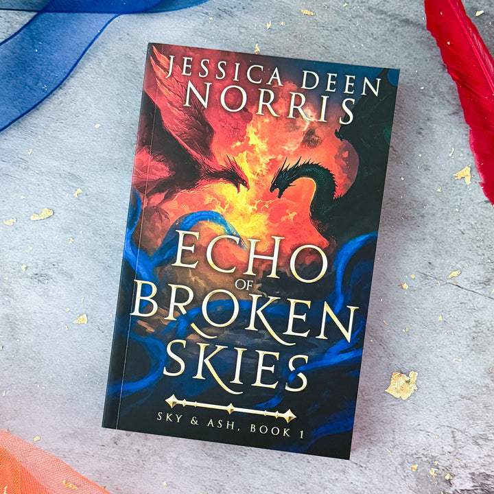 Paperback - Echo of Broken Skies by Jessica Deen Norris - Cover features the author's name at the top in large serif font. The central artwork depicts a dramatic sky divided by swirling clouds of fire. On the left, a red phoenix with outstretched wings flies toward a dark green dragon on the right, both poised as if mid-battle. Blue, smoky tendrils swirl around the bottom half of the image. The title is centered in bold, metallic gold, with "Sky & Ash, Book 1" below a decorative gold divider.