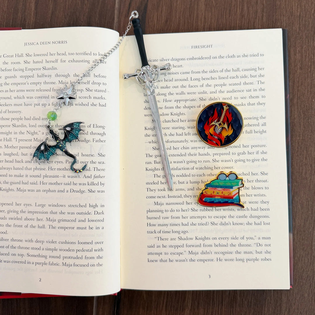 Sword bookmark with a chain and dragon charm hanging from it, plus two enamel pins. One pin features a sword with flames surrounding it and the other is a stack of books with glasses on top and a dragon tail wrapping around the stack.