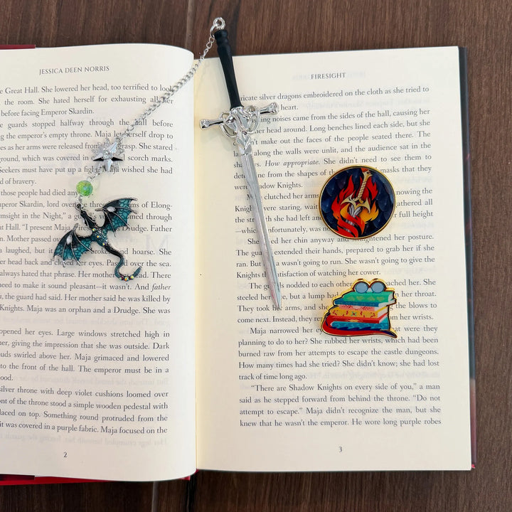 Sword bookmark with a chain and dragon charm hanging from it, plus two enamel pins. One pin features a sword with flames surrounding it and the other is a stack of books with glasses on top and a dragon tail wrapping around the stack.