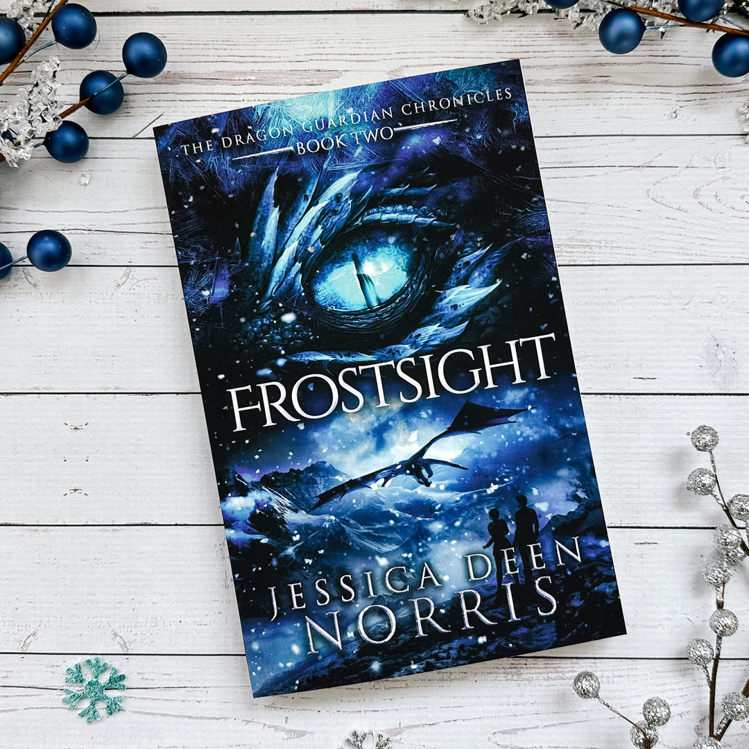 Frostsight paperback cover. Cover features the title across the middle with a blue dragon eye and "The Dragon Guardian Chronicles Book 2" above it. Below the title, the two main characters watch a dragon in the distance in a snowy mountain landscape. The author's name is at the bottom of the cover.