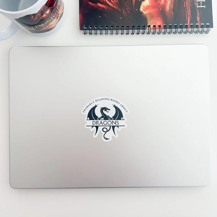 A white vinyl sticker on a laptop with a black design that reads "Probably reading books about dragons." The word "dragons" is set apart on top of a large dragon.