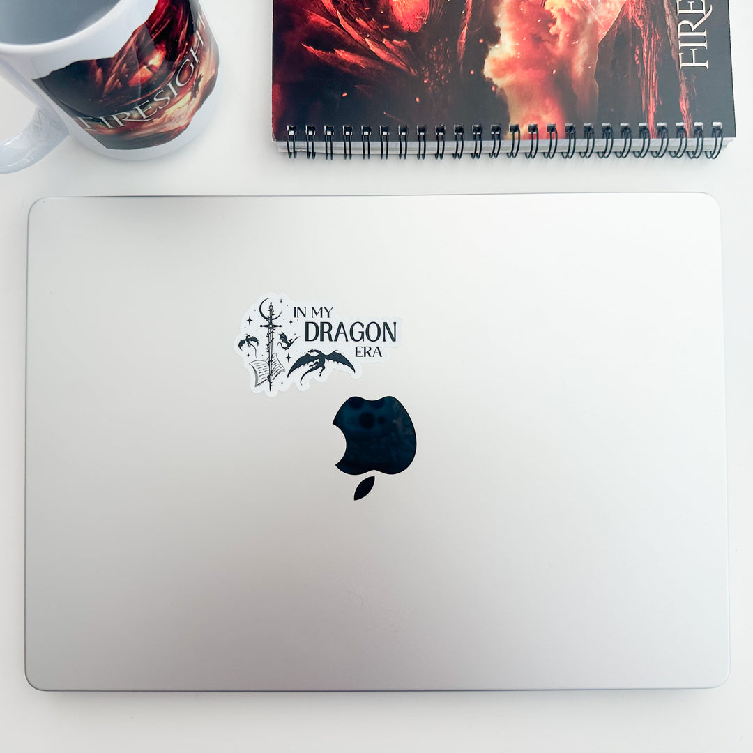 A vinyl sticker on a laptop that reads "In my dragon era." Next to the text are a sword, a book, magical stars and moon, and flying dragons.