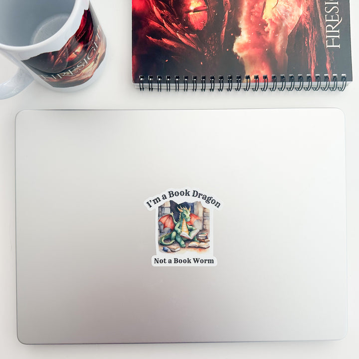 A vinyl sticker on a laptop that reads "I'm a book dragon not a book worm." Between the text is a watercolor design of a dragon reading next to a stack of books.