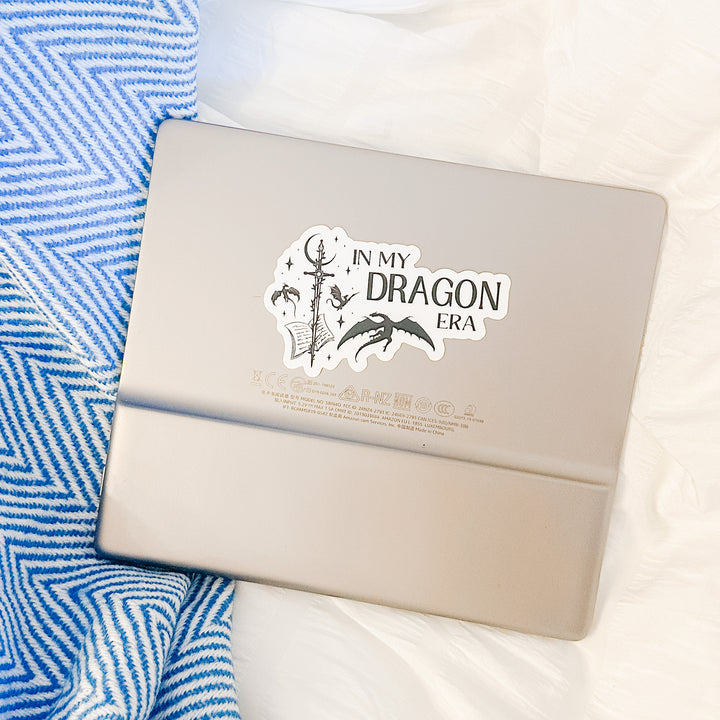 A vinyl sticker on a Kindle that reads "In my dragon era." Next to the text are a sword, a book, magical stars and moon, and flying dragons.