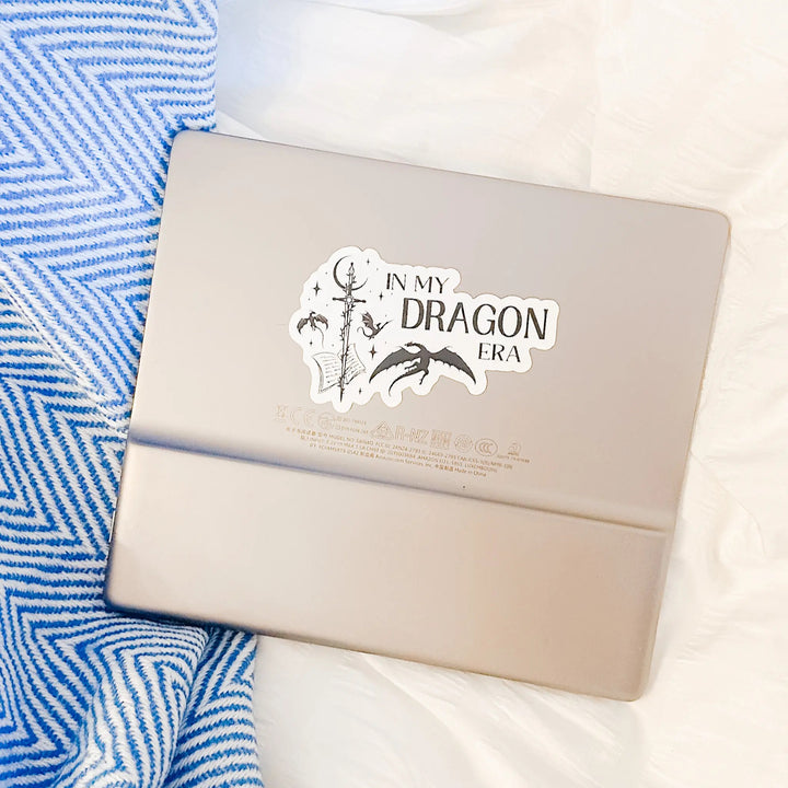 A vinyl sticker on a Kindle that reads "In my dragon era." Next to the text are a sword, a book, magical stars and moon, and flying dragons.