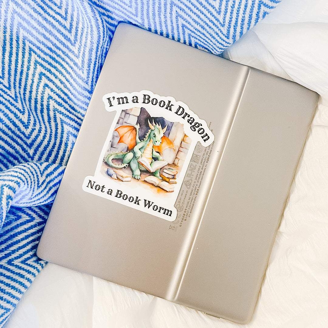 A vinyl sticker on a Kindle that reads "I'm a book dragon not a book worm." Between the text is a watercolor design of a dragon reading next to a stack of books.