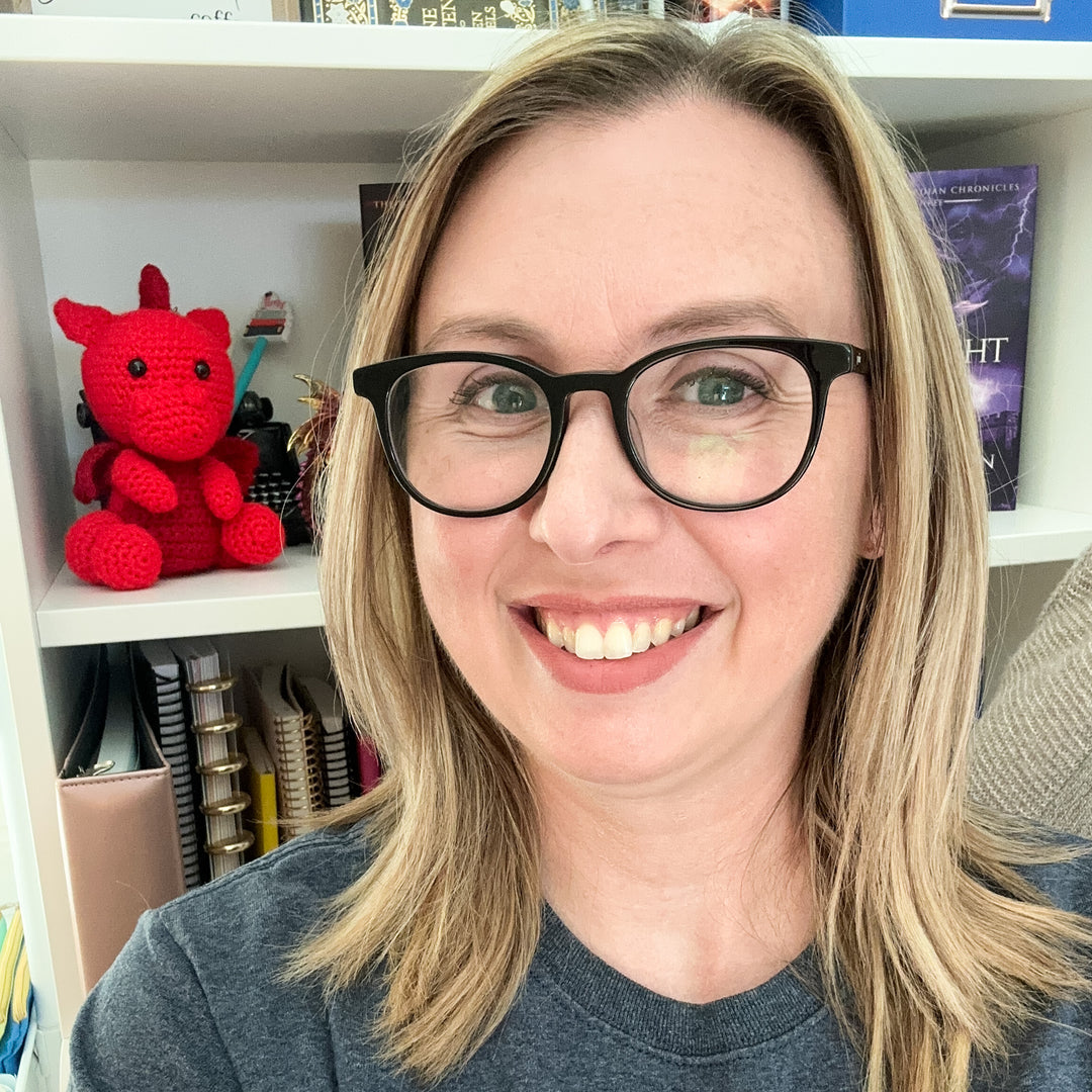 Jessica Deen Norris smiling in front of her bookshelf. A small red crocheted dragon sits on the shelf behind her.