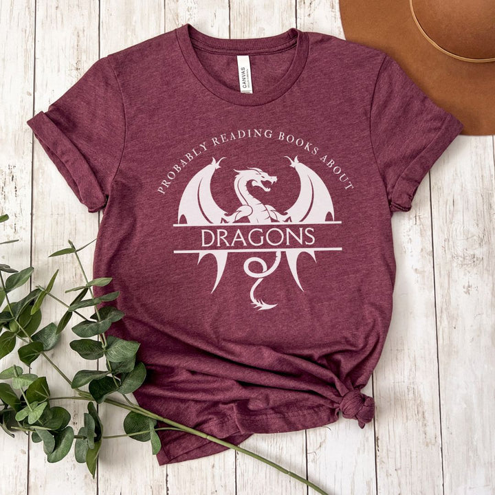 A short sleeve heather maroon t-shirt. The white design on the shirt reads "Probably reading books about dragons." The word "dragons" is set apart on top of a large dragon.