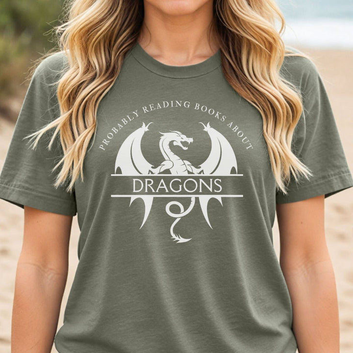 A woman wearing a short sleeve heather military green t-shirt. The white design on the shirt reads "Probably reading books about dragons." The word "dragons" is set apart on top of a large dragon.