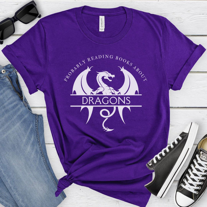 A short sleeve purple t-shirt. The white design on the shirt reads "Probably reading books about dragons." The word "dragons" is set apart on top of a large dragon.