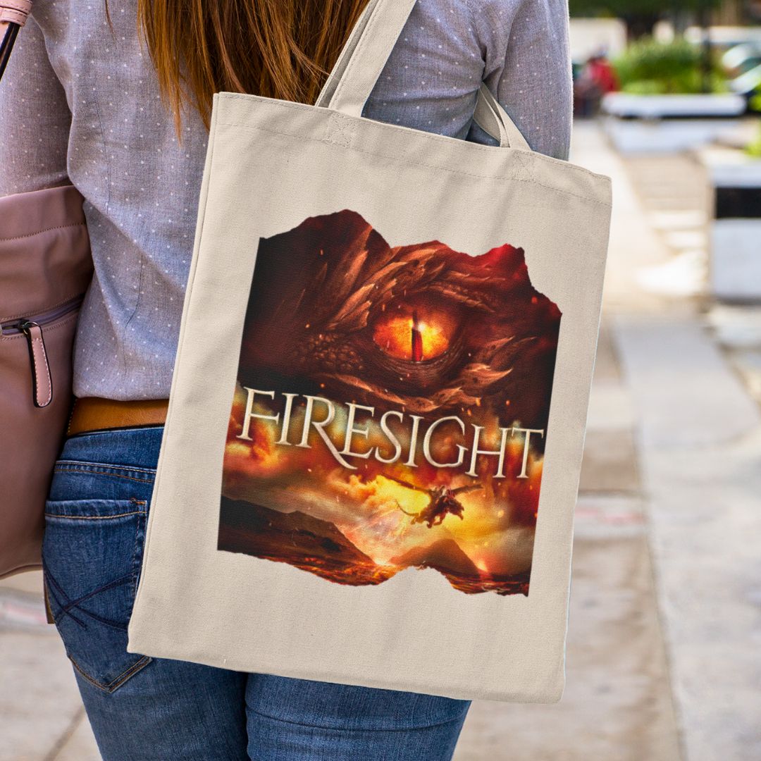 Someone holding a tan canvas tote bag. Design on the tote bag is the cover of Firesight, just showing the red dragon eye and the title with the two main characters riding on a dragon over a volcano erupting.