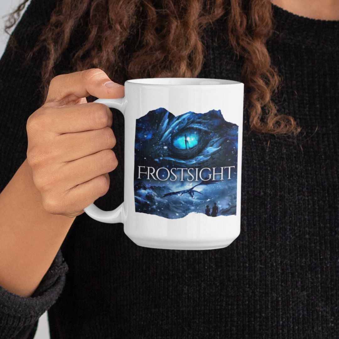Someone holding a white mug. Design on the mug is the cover of Frostsight, just showing the blue dragon eye and the title with the two main characters watching the dragon in the snowy mountain landscape.