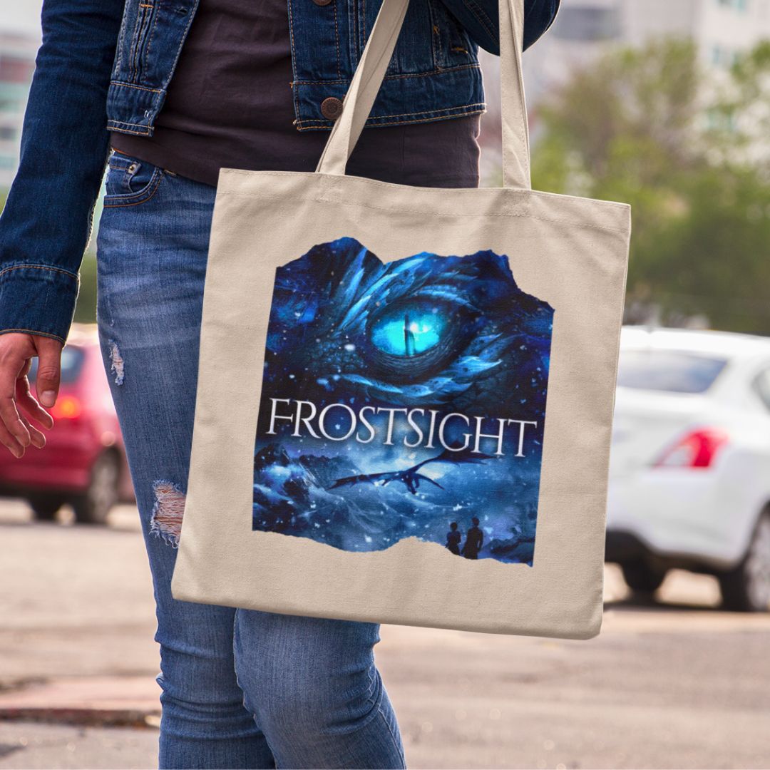 Someone holding a tan canvas tote bag. Design on the tote bag is the cover of Frostsight, just showing the blue dragon eye and the title with the two main characters watching the dragon in the snowy mountain landscape.